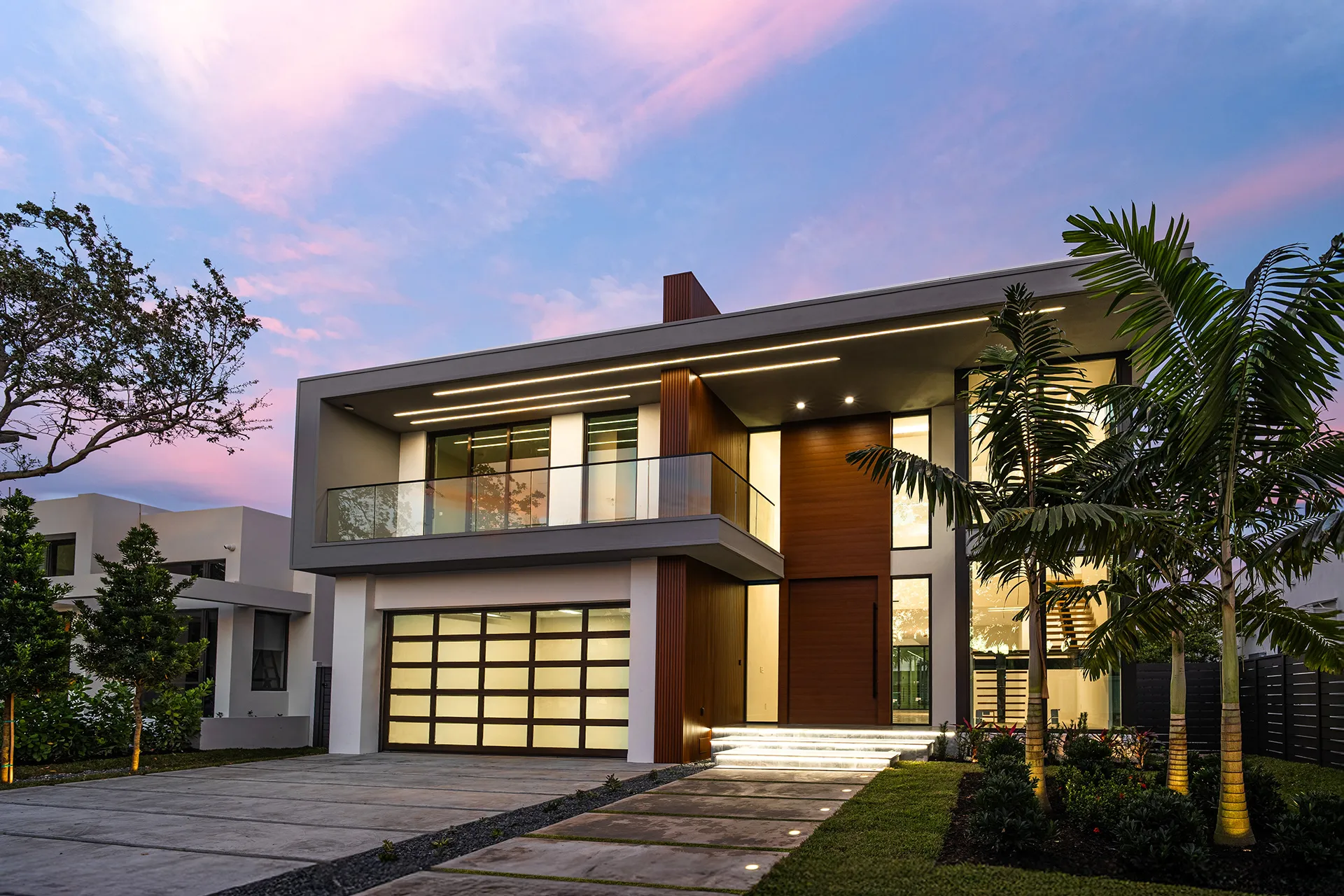 Modern two-storey home at dusk, warm interior light and palm trees
